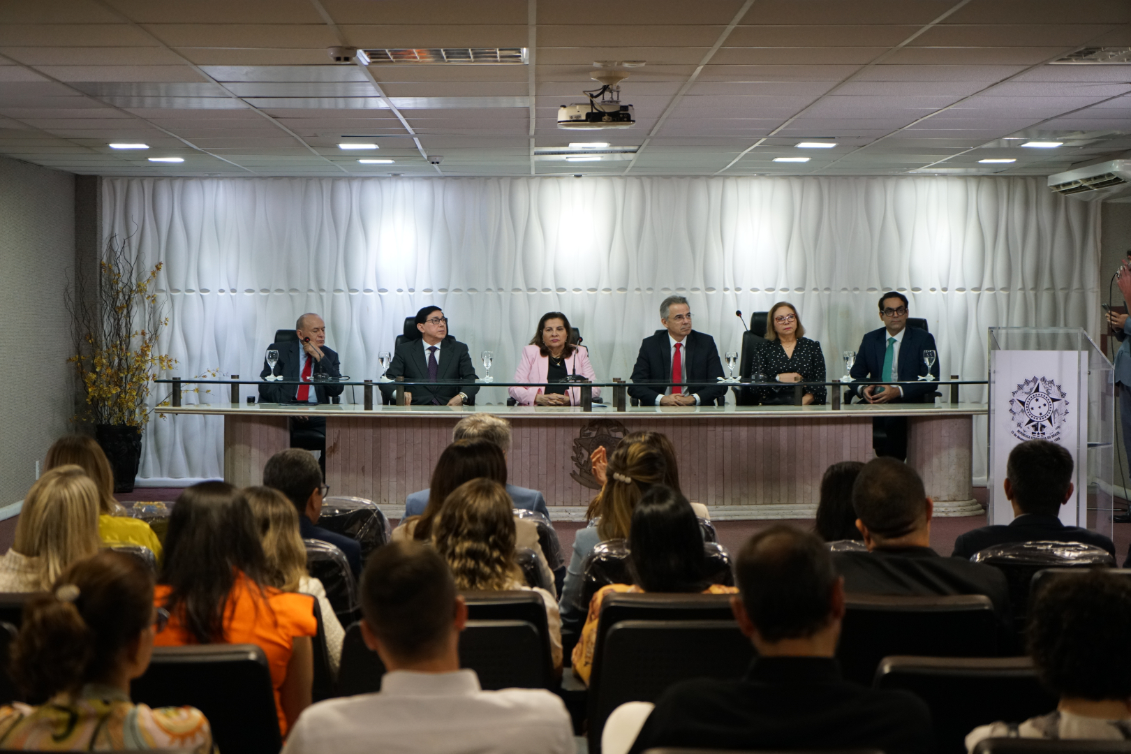  Sete pessoas, incluindo uma mulher no centro segurando um documento, posam em fila. Todos vestem trajes formais em um ambiente interno bem iluminado. A imagem captura um momento de formalidade e colaboração.