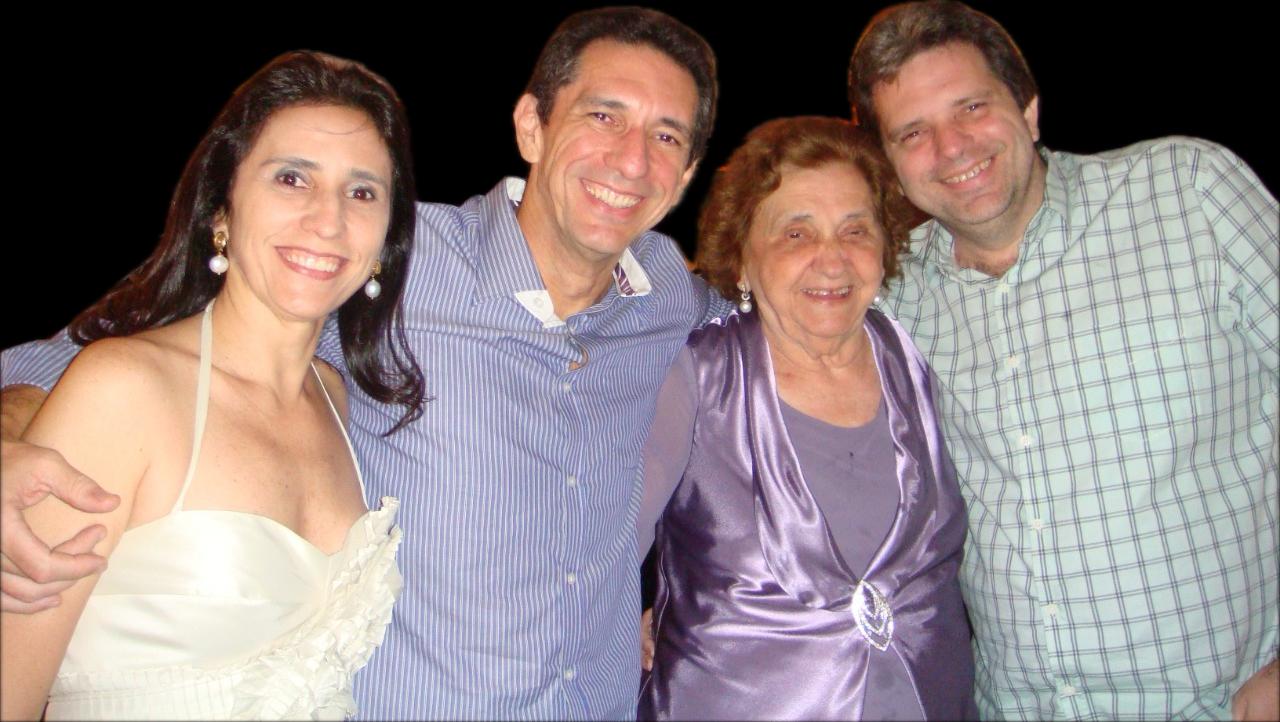 Quatro pessoas sorriem para a câmera em um fundo escuro. Uma mulher de vestido branco, dois homens com camisas diferentes e uma senhora de roxo posam juntos. Representam dona Zilca e seus três filhos