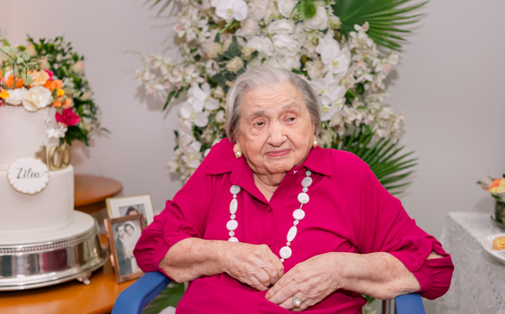 Uma senhora idosa de cabelos brancos e blusa magenta está sentada em uma cadeira de rodas azul. Ao fundo, há arranjos florais brancos e um bolo de aniversário branco com flores coloridas. Ela segura as mãos no colo e observa algo à direita.