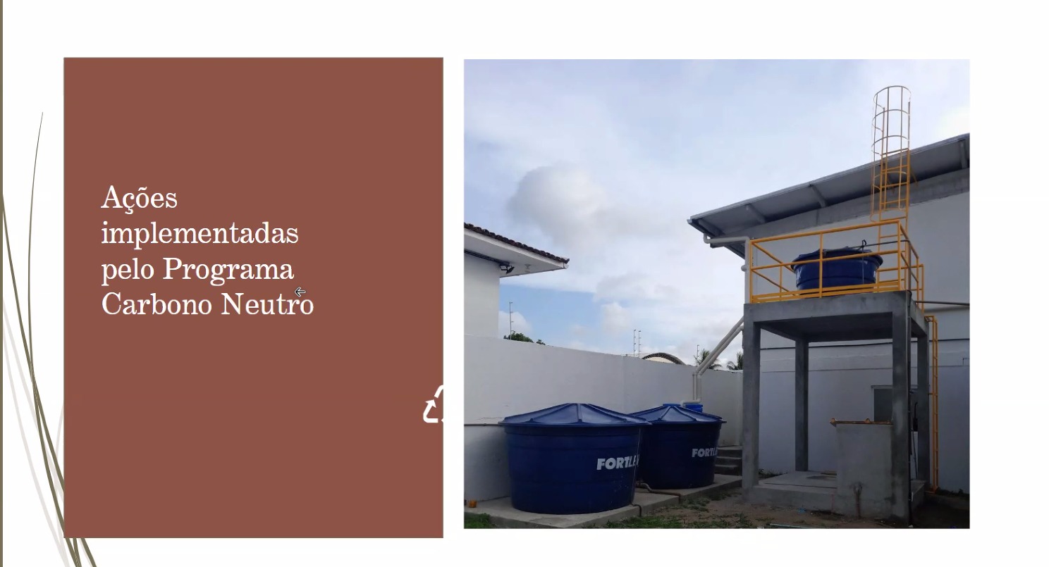  Uma tela de apresentação exibe um slide dividido em duas partes. O lado esquerdo tem um fundo marrom escuro com o texto em branco: "Ações implementadas pelo Programa Carbono Neutro". O lado direito mostra uma fotografia externa de um edifício. Na imagem, há dois grandes reservatórios de água azuis no nível do chão e um terceiro reservatório azul menor elevado em uma estrutura de concreto com uma escada amarela de acesso. A parte superior do edifício tem um telhado de telhas e um calha. O céu está parcialmente nublado.
