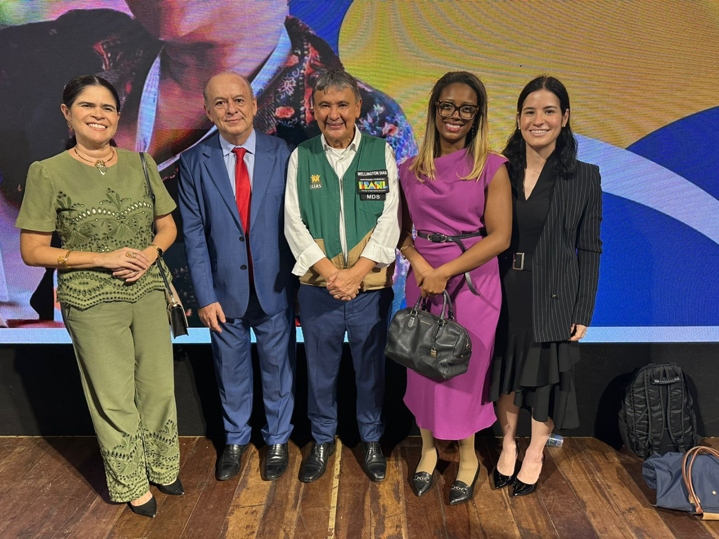 Cinco pessoas, três mulheres e dois homens, posam lado a lado em frente a um telão vibrante. A mulher à esquerda veste um conjunto verde-oliva com bordados; o homem ao lado dela usa um terno azul e gravata vermelha. No centro, um homem de colete verde sobre camisa branca; à sua direita, uma mulher de vestido roxo segura uma bolsa preta, e por último, uma mulher de preto com um blazer risca de giz.