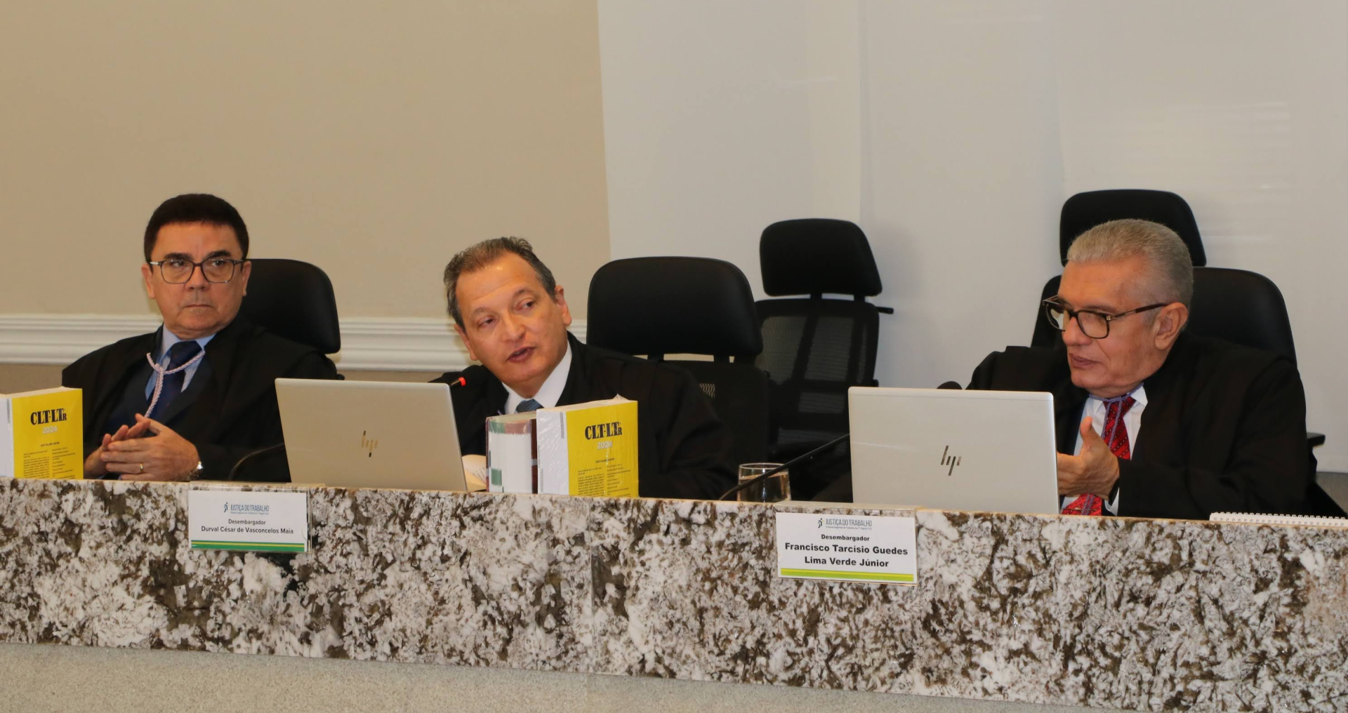  A imagem apresenta três homens de meia-idade, sentados em uma mesa de mármore. O homem do centro está falando (Des. Durval Maia), enquanto os outros dois o escutam. Todos vestem ternos escuros, e há laptops à frente de cada um. Placas de identificação sobre a mesa indicam os nomes.