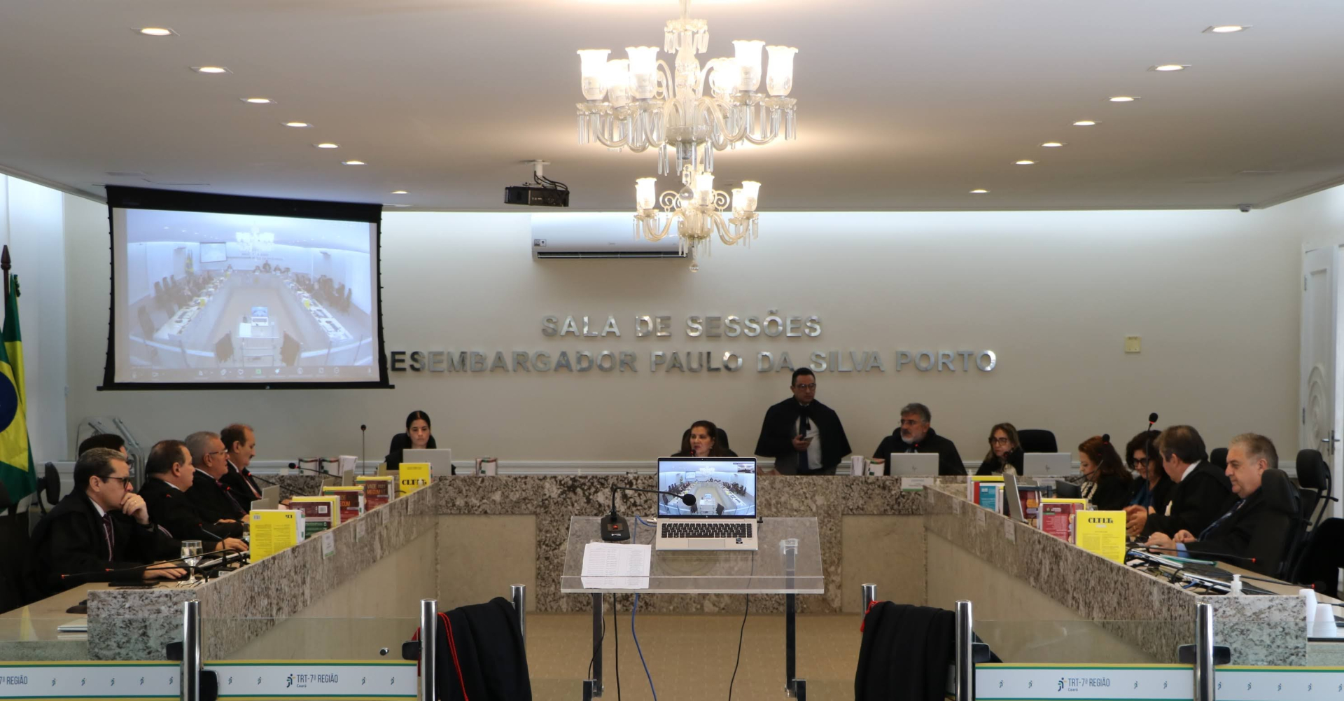  A imagem mostra a "SALA DE SESSÕES DESEMBARGADOR PAULO DA SILVA PORTO", com uma grande mesa retangular em formato de "U" onde estão sentados diversos magistrados de toga preta. Ao fundo, um telão exibe uma videochamada, e um lustre imponente pende do teto. No centro da mesa, um laptop com a mesma imagem do telão é visível