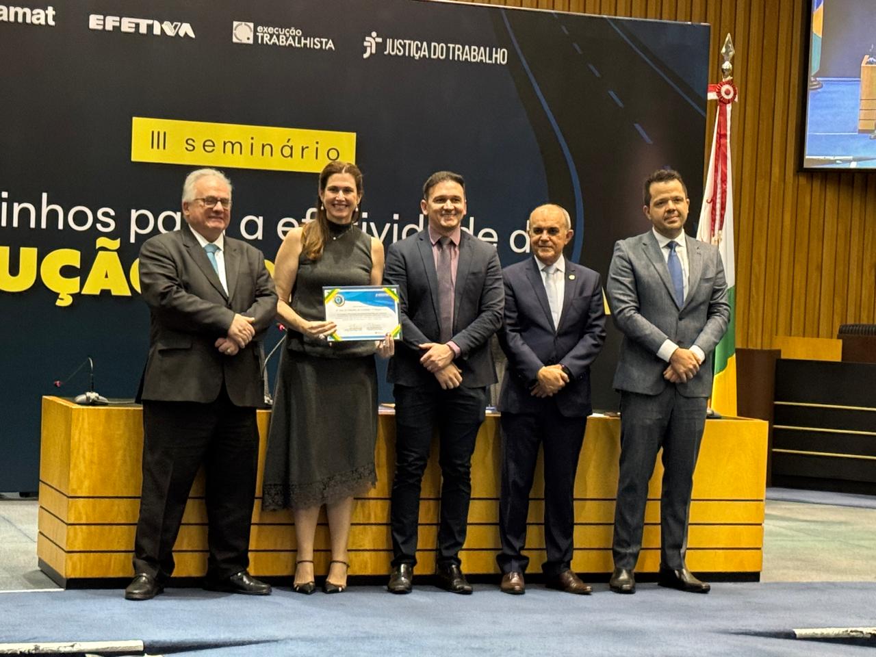  No dia 10 de junho de 2025, em um auditório em Fortaleza, Ceará, cinco pessoas, quatro homens de terno e uma mulher de vestido escuro, estão em pé em um palco. A mulher segura um certificado de cor azul e branca. Ao fundo, um grande painel preto exibe logotipos de instituições como "EXECUÇÃO TRABALHISTA" e "JUSTIÇA DO TRABALHO", com o texto "III seminário" e uma frase parcial.