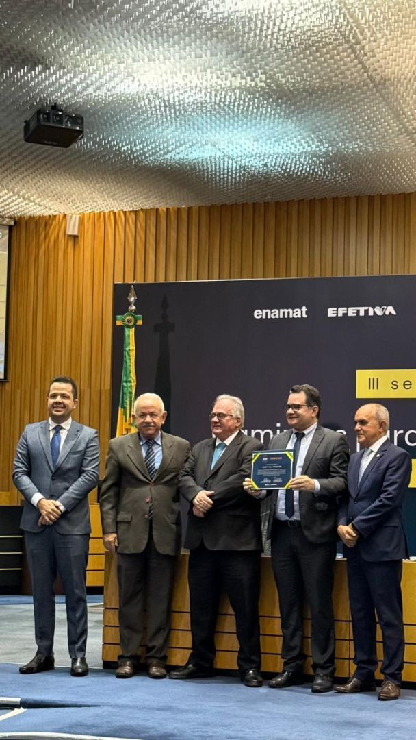  Um grupo de cinco homens de terno está em pé em um palco, com um deles segurando um certificado. Ao fundo, há um painel preto com logotipos e a palavra "seminário", além de uma bandeira do Brasil. O ambiente é um auditório com paredes de madeira.