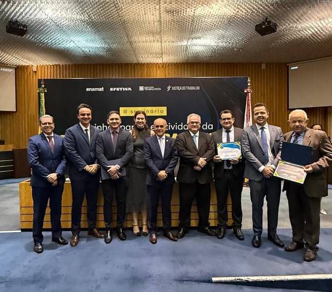 Em um auditório, um grupo de nove pessoas, incluindo oito homens de terno e uma mulher de vestido escuro, posa para uma foto. Dois dos homens seguram certificados de cor azul. Ao fundo, um painel preto exibe logotipos de instituições relacionadas à Justiça do Trabalho