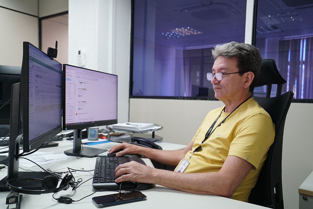 Homem de pele clara, óculos de grau e camiseta amarela trabalha sentado em uma cadeira de escritório preta. Ele está focado, com as duas mãos operando o teclado e o mouse em uma mesa com dois monitores ligados. Ao fundo, uma grande divisória de vidro reflete o ambiente interno e exibe uma janela ao fundo da sala.
