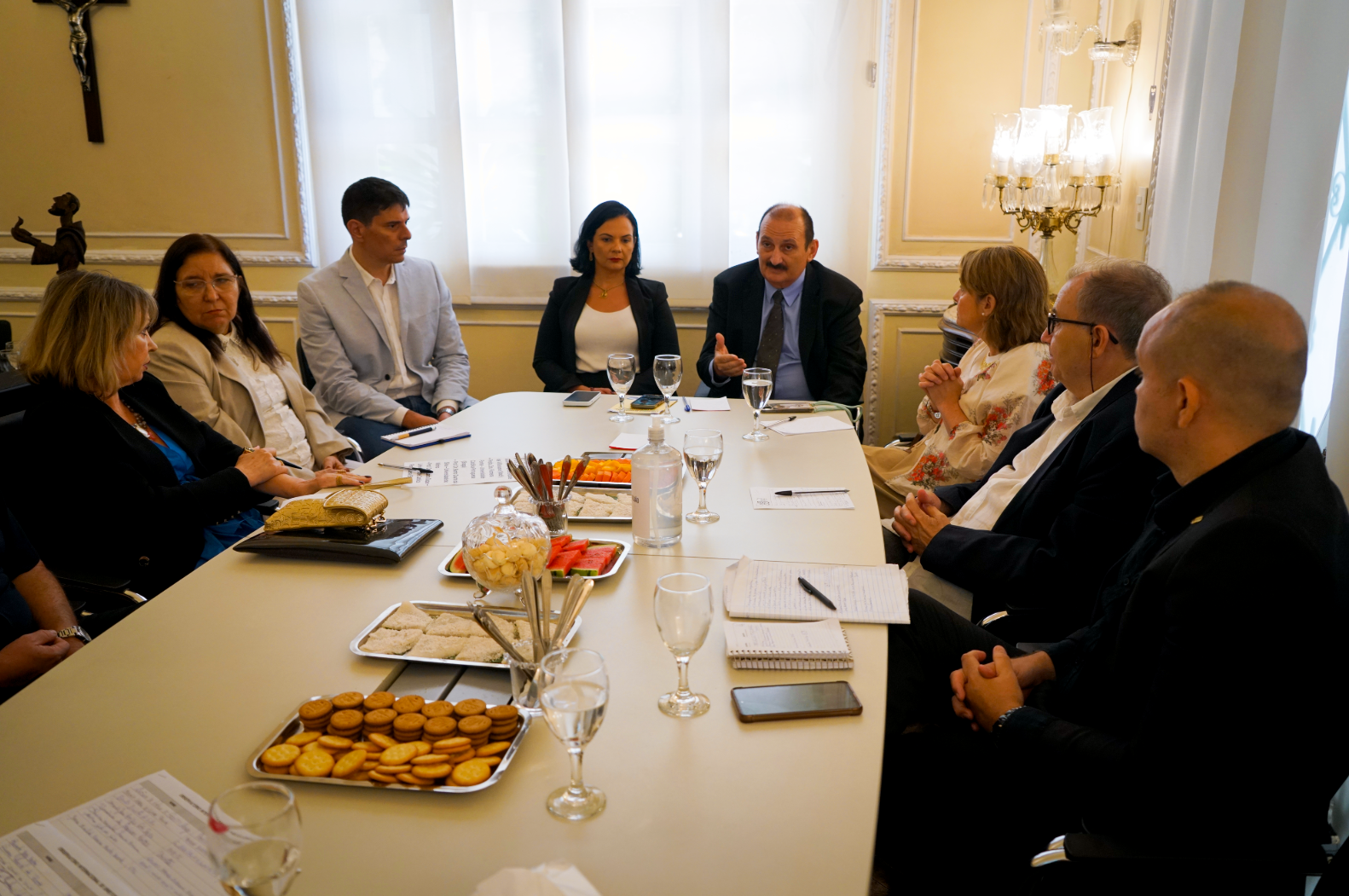 A imagem retrata um grupo de nove pessoas sentadas ao redor de uma mesa retangular clara, em um ambiente formal que parece ser uma sala de reuniões. Há alguns homens e mulheres, predominantemente adultos, vestidos em trajes sociais ou esporte fino, conversando e alguns comendo petiscos como biscoitos e frutas dispostos em bandejas. Copos com água ou vinho, blocos de anotações e canetas também estão sobre a mesa. Ao fundo, cortinas claras e uma estante com um crucifixo na parede.