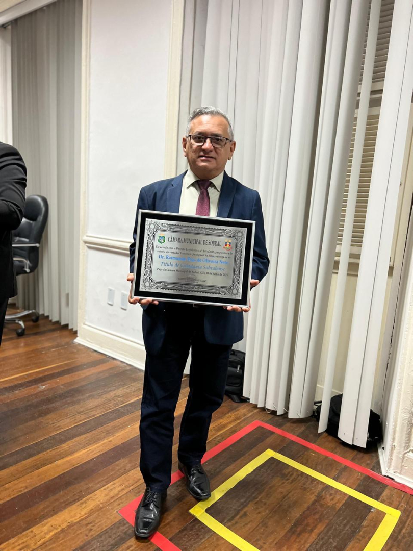 Um homem de óculos e terno escuro segura uma placa prateada com a inscrição "Câmara Municipal de Sobral". Ele está em pé sobre um piso de madeira com detalhes em vermelho e amarelo, e ao fundo há uma parede com cortinas brancas.