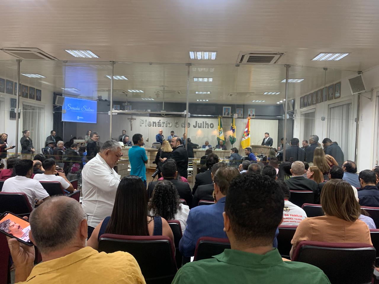 Uma visão ampla de um plenário cheio, com diversas pessoas sentadas em cadeiras escuras, observando a parte frontal do ambiente. Ao fundo, um púlpito e uma mesa onde autoridades estão sentadas, com a faixa "Plenário 5 de Julho" visível na parede. Há um telão à esquerda exibindo "Sessão Solene".