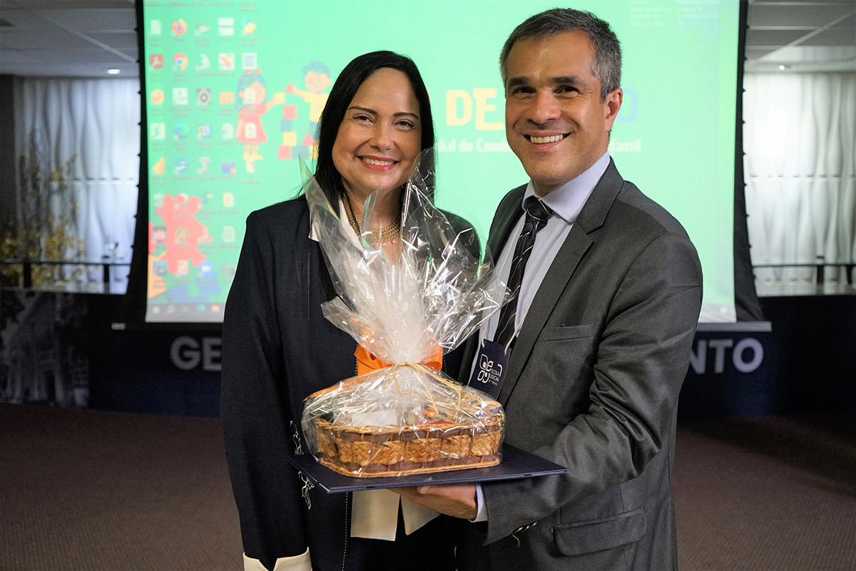 Um homem sorridente de terno e uma mulher com um blazer escuro, também sorridente, posam juntos. O homem segura um presente embrulhado em celofane, exibindo seu conteúdo semelhante a doces ou pães. Ao fundo, uma tela projeta uma imagem colorida.