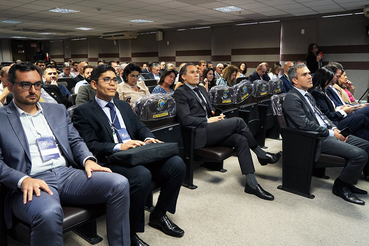 Um auditório com várias pessoas sentadas em cadeiras acolchoadas, prestando atenção ao que acontece à frente. Os assentos são de couro e as pessoas estão vestidas predominantemente em trajes formais, como ternos.