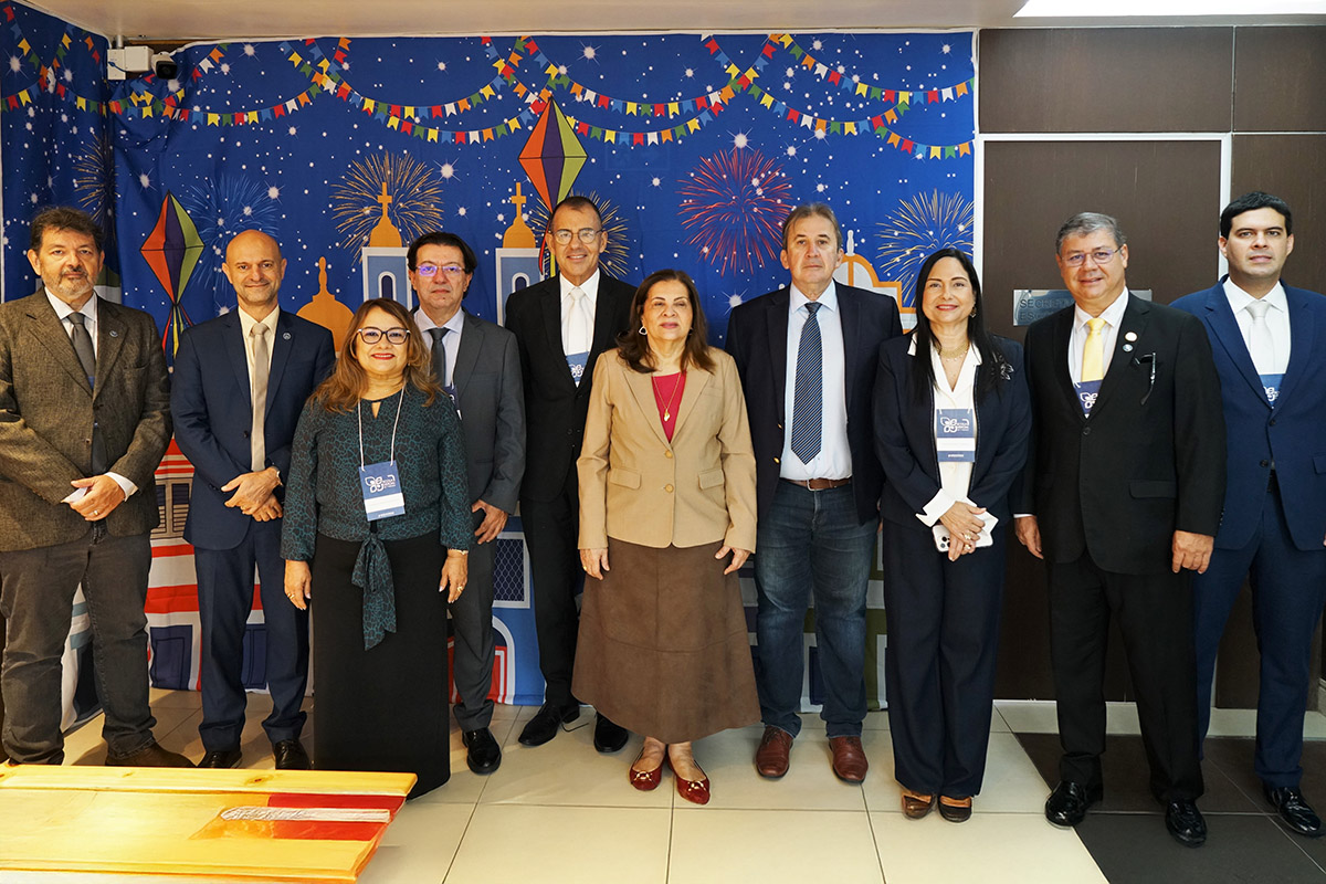 Dez pessoas posam em pé para uma foto em grupo, em frente a uma parede decorada com motivos de festa junina, incluindo balões e fogos de artifício. Todos estão vestidos formalmente, com alguns usando crachás.