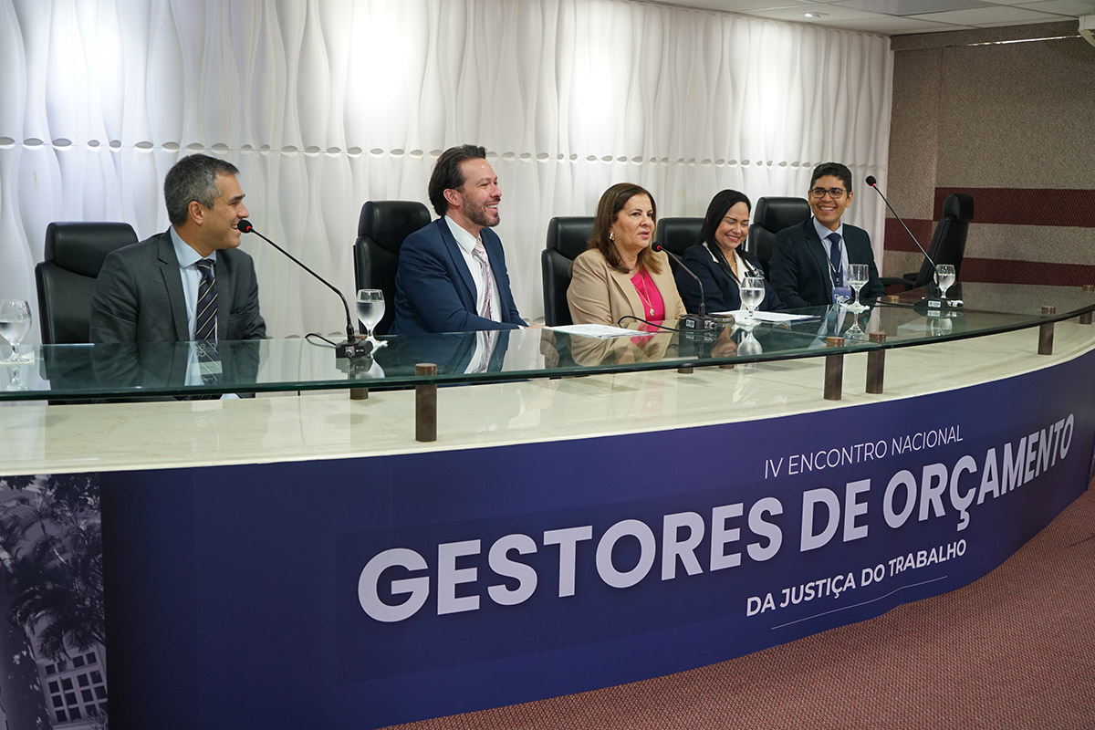 Cinco pessoas estão sentadas em uma mesa de conferência com microfones e copos d'água. Quatro homens e uma mulher parecem engajados em uma discussão ou apresentação, com um banner azul à frente da mesa que diz "IV ENCONTRO NACIONAL GESTORES DE ORÇAMENTO DA JUSTIÇA DO TRABALHO".