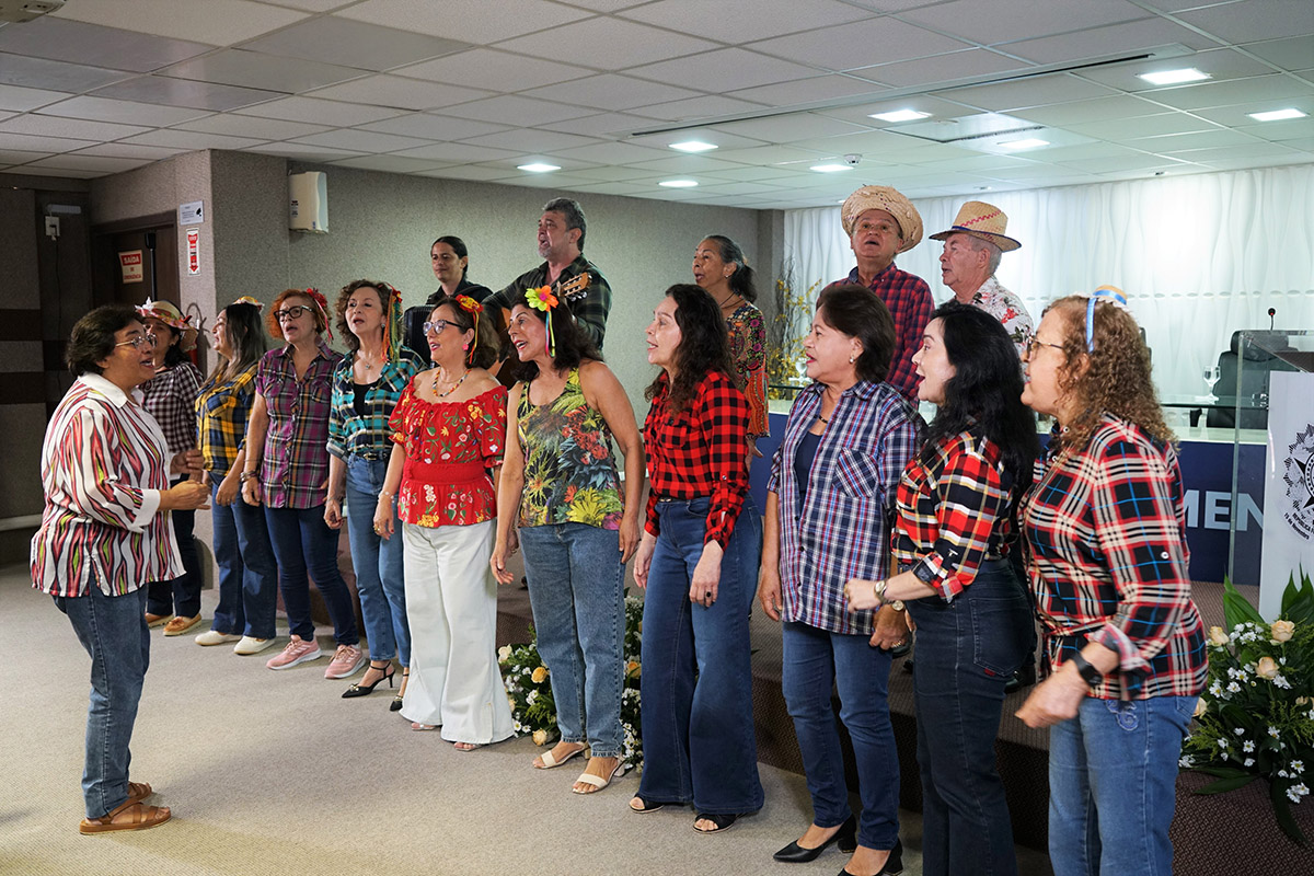 Um grande grupo de pessoas, a maioria mulheres, canta ou se apresenta em um evento interno. Muitos usam camisas xadrez, enquanto alguns homens ao fundo usam chapéus de palha, sugerindo um evento de festa junina.