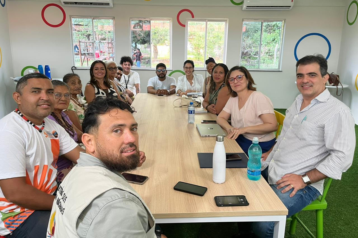 Uma sala clara com decoração de círculos coloridos nas paredes. Um grupo de cerca de 15 pessoas, incluindo homens e mulheres de várias idades, está sentado ao redor de uma longa mesa de madeira. Todos sorriem para a câmera, que os enquadra do lado esquerdo da mesa.