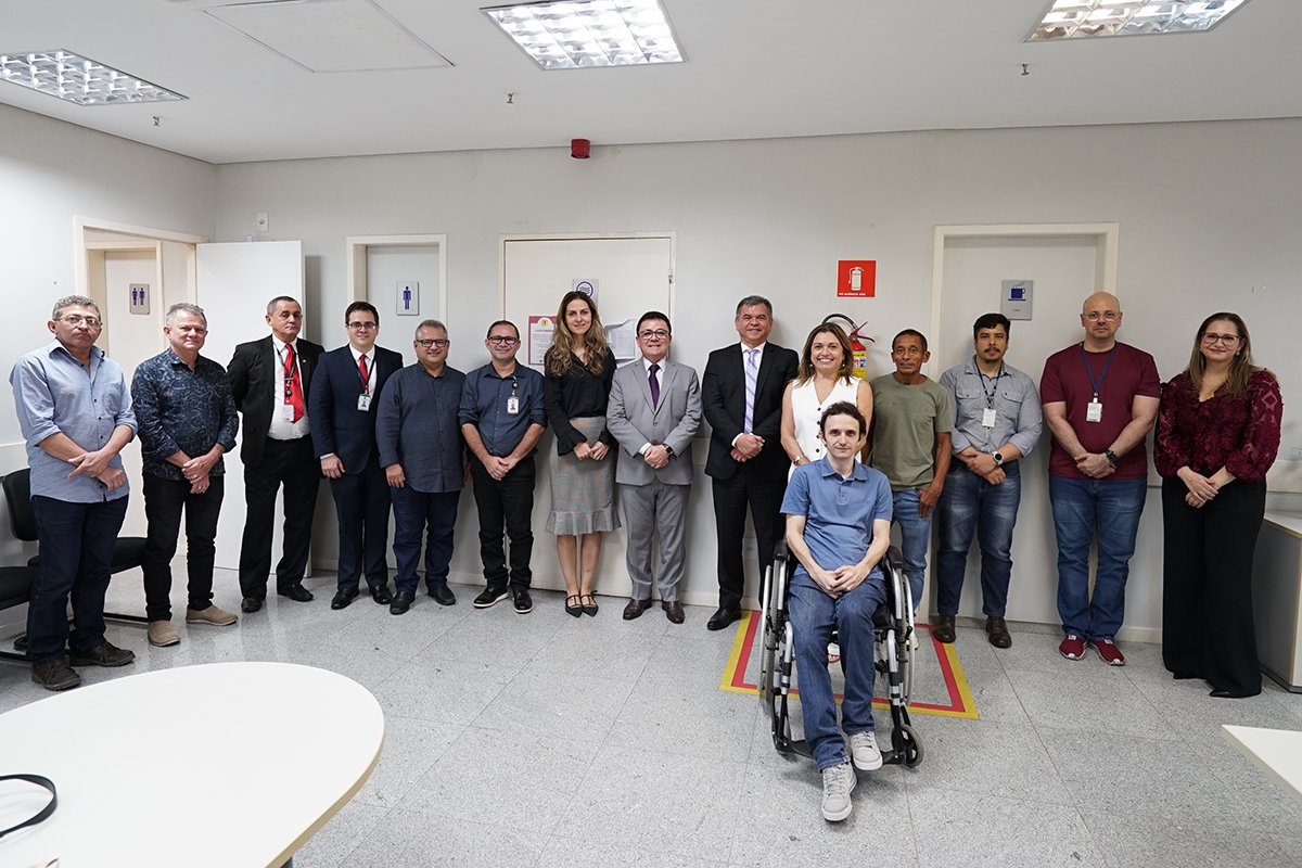 Um grupo de dezesseis pessoas está em uma sala de escritório, com paredes brancas e iluminação de teto. Um homem em cadeira de rodas está sentado na frente do grupo, olhando para a câmera e sorrindo. As outras pessoas estão em pé atrás dele, formando uma fila, algumas sorrindo.