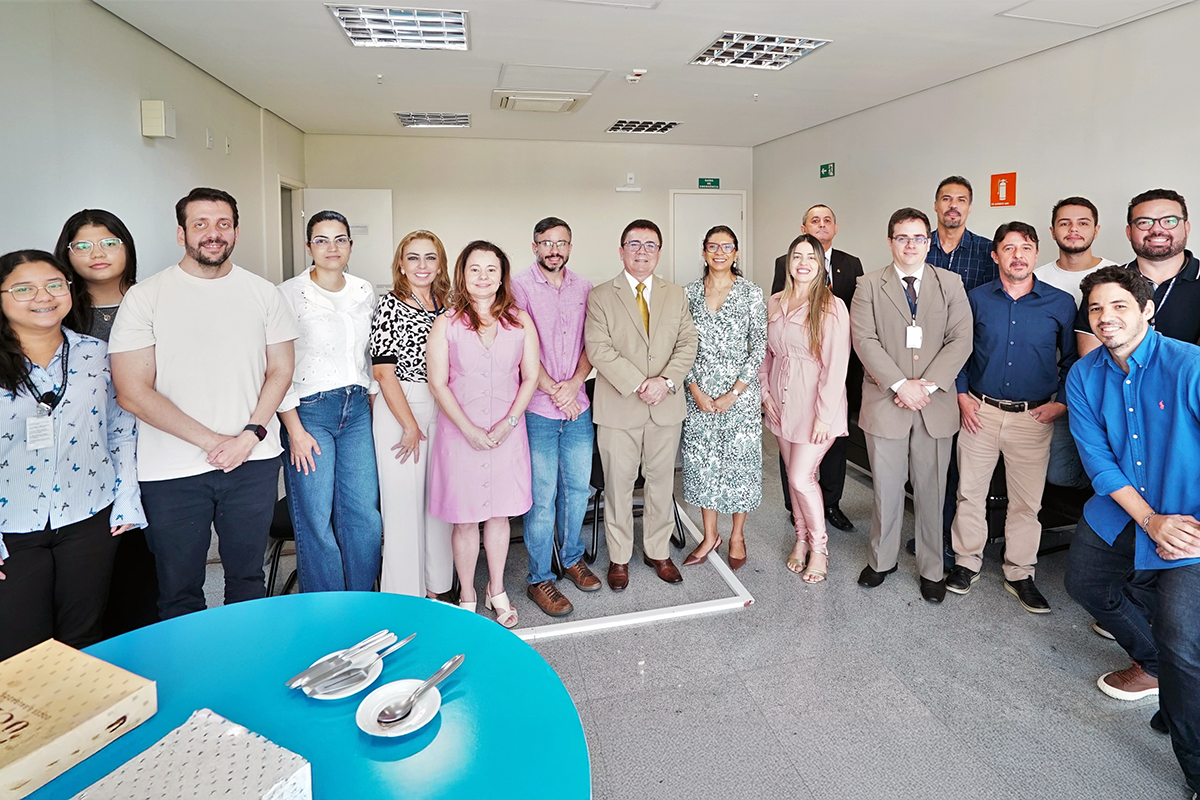 Fotografia de um grupo de quinze pessoas em pé, posando em um ambiente de escritório claro e moderno. No centro, um homem de terno bege e óculos é o ponto focal, cercado por colegas. As vestimentas variam entre o social e o casual. No primeiro plano, uma mesa redonda azul com uma caixa e talheres sobre ela compõe a cena.