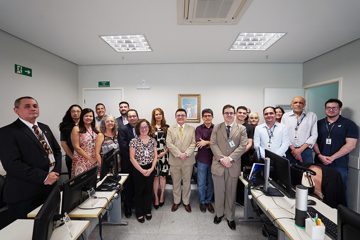 Fotografia de um grupo de quinze pessoas em um escritório. Estão todos de pé, lado a lado, posando para a foto em um ambiente com mesas de trabalho e computadores. Ao centro, um homem de terno bege e óculos sorri, ladeado por homens e mulheres em trajes sociais e esporte fino. A iluminação vem do teto e a atmosfera é profissional.