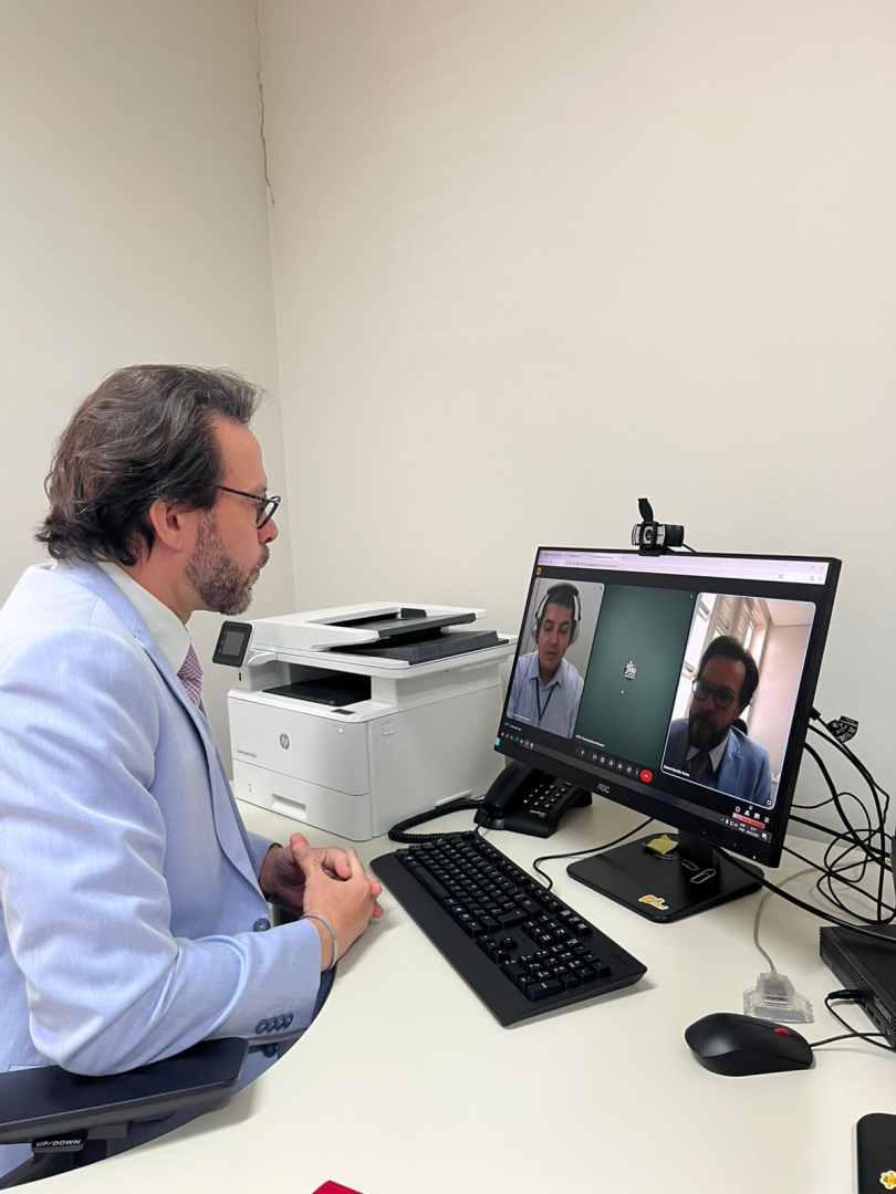 Homem de óculos e terno azul claro está sentado em uma mesa branca, de perfil para a esquerda, olhando para uma tela de computador onde duas pessoas participam de uma videochamada. Ao lado do monitor, há uma impressora branca.