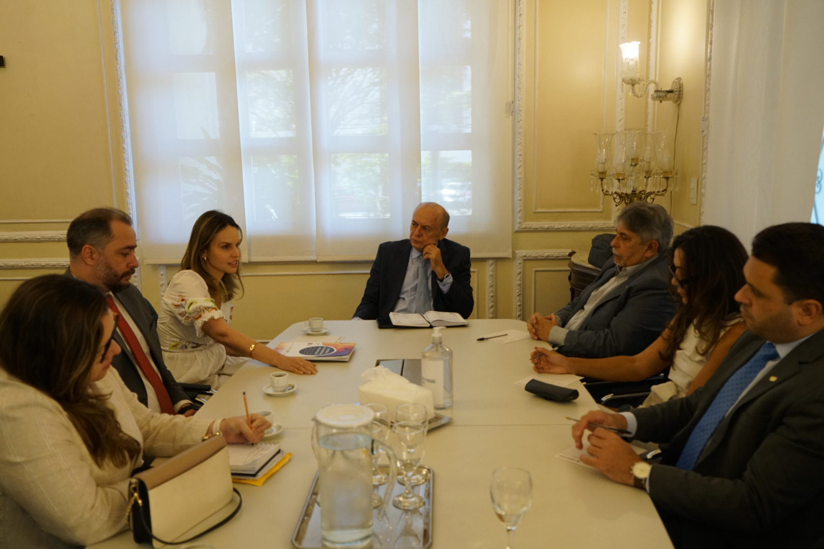 Uma foto colorida mostra sete pessoas sentadas ao redor de uma mesa de reunião retangular. Eles estão conversando e interagindo, com papéis, xícaras de café e uma jarra de água sobre a mesa. A sala tem uma grande janela com persianas ao fundo e uma decoração clássica com luminárias nas paredes.