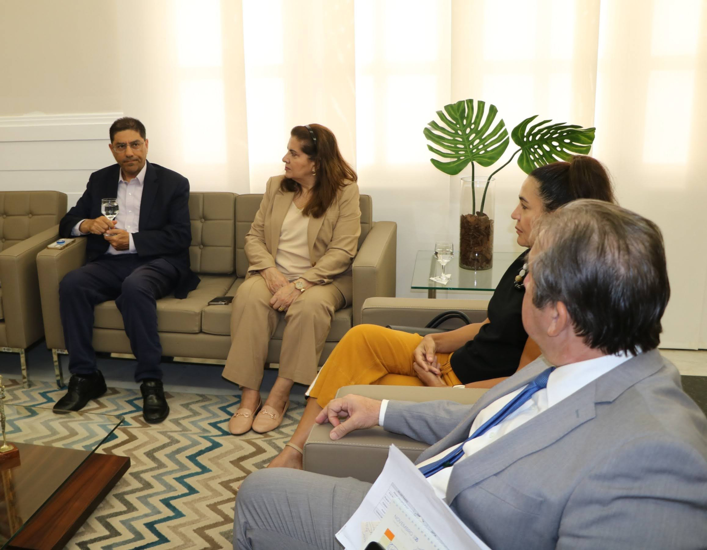 Um homem de terno cinza e uma mulher de blazer branco estão sentados à mesa, com microfones à frente. Ambos sorriem enquanto olham para a câmera. Ao fundo, um ambiente de escritório com uma janela.