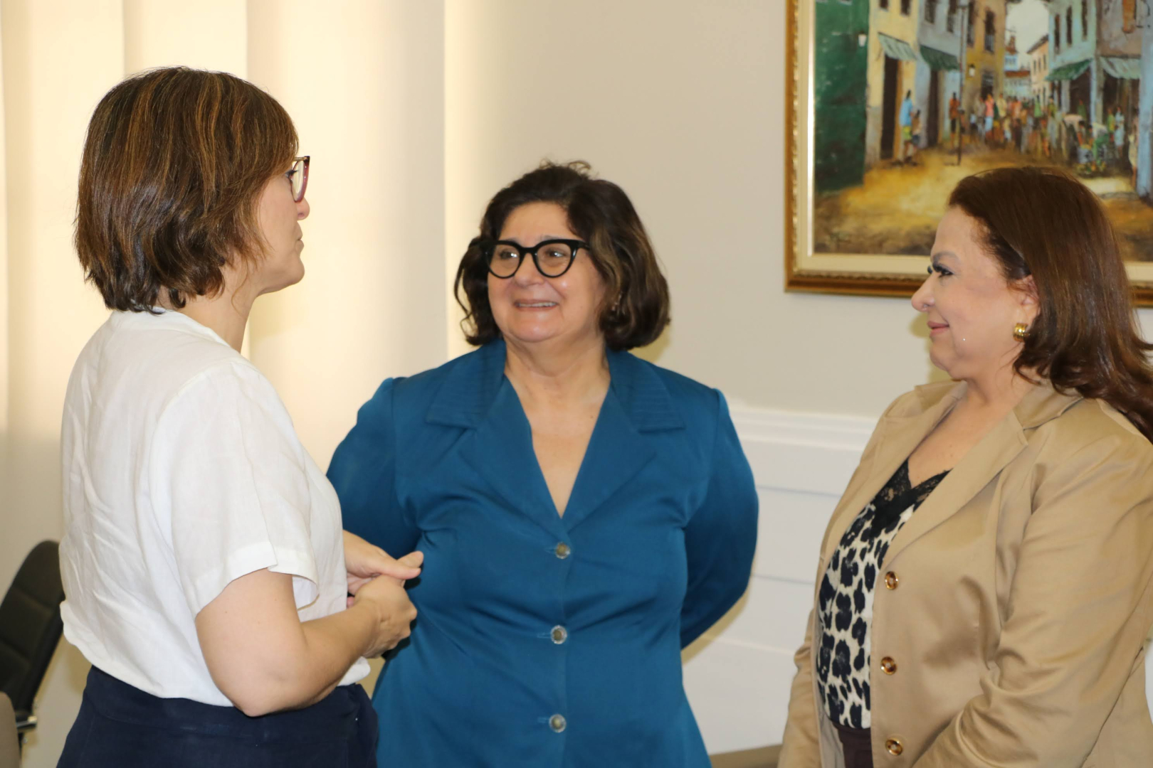 Três mulheres, uma de blusa branca e as outras duas de blazer, conversam sorridentes em um ambiente interno. A mulher do centro usa óculos e blazer azul, enquanto a da direita veste blazer bege e blusa estampada. Ao fundo, um quadro com paisagem.