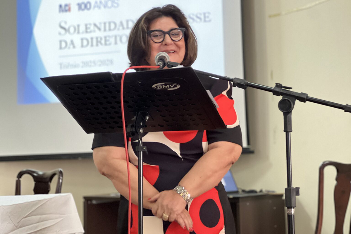 Uma mulher de óculos e vestido estampado preto, vermelho e branco está em pé atrás de um púlpito com um microfone, falando para um público. Ao fundo, um telão branco exibe o texto "ACI 100 ANOS SOLENIDADE DA DIRETORIA".