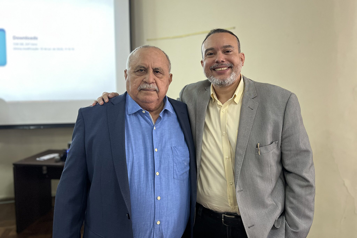 Dois homens posam para a foto com um braço no ombro um do outro. O homem à esquerda usa uma camisa azul e o da direita uma camisa social clara com um blazer cinza. Ambos sorriem.