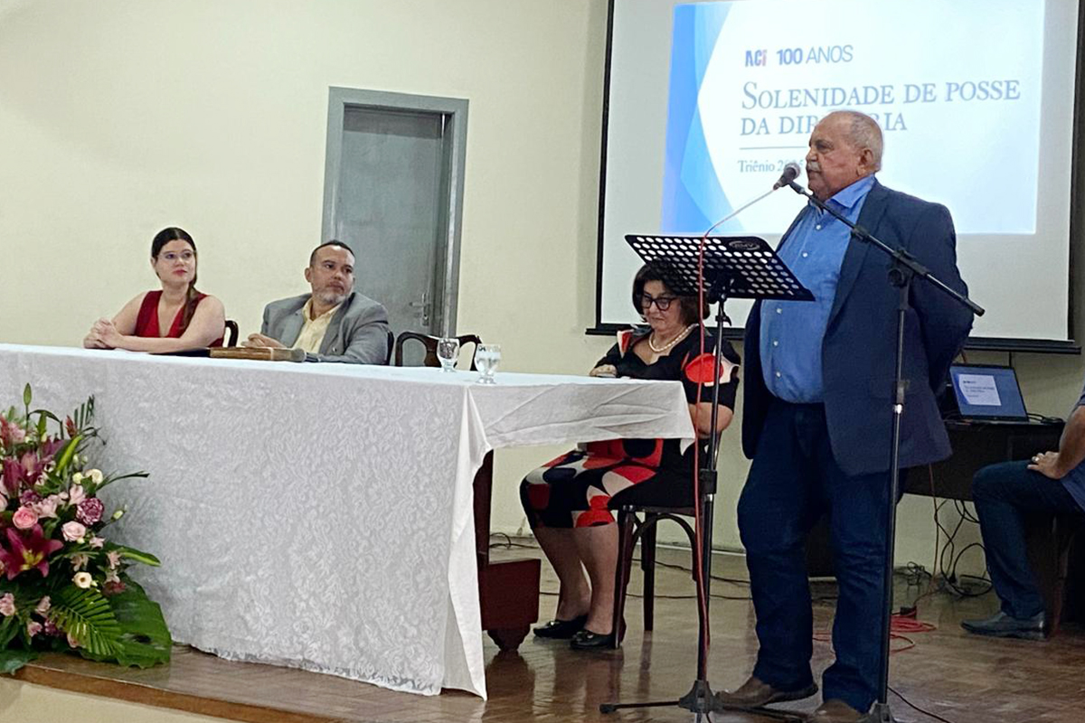 Um homem de camisa azul está em pé no palco, falando ao microfone, com um púlpito à sua frente. À mesa, sentados, estão uma mulher de blusa vermelha, um homem de blazer cinza e uma mulher com vestido estampado. Um telão ao fundo exibe o texto "ACI 100 ANOS SOLENIDADE DE POSSE DA DIRETORIA".