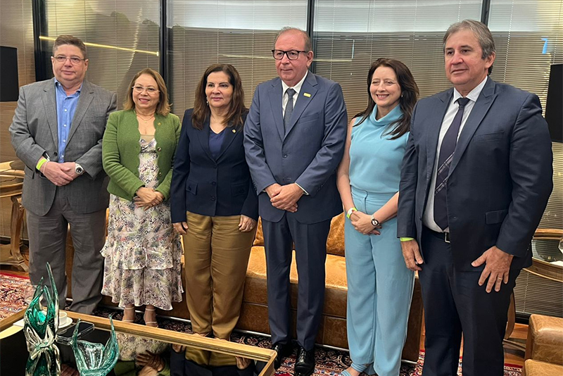  Cinco pessoas estão em pé em um ambiente interno. Da esquerda para a direita, há dois homens e três mulheres. O homem do centro veste um terno azul-marinho. Todos estão bem vestidos e olham para a frente.