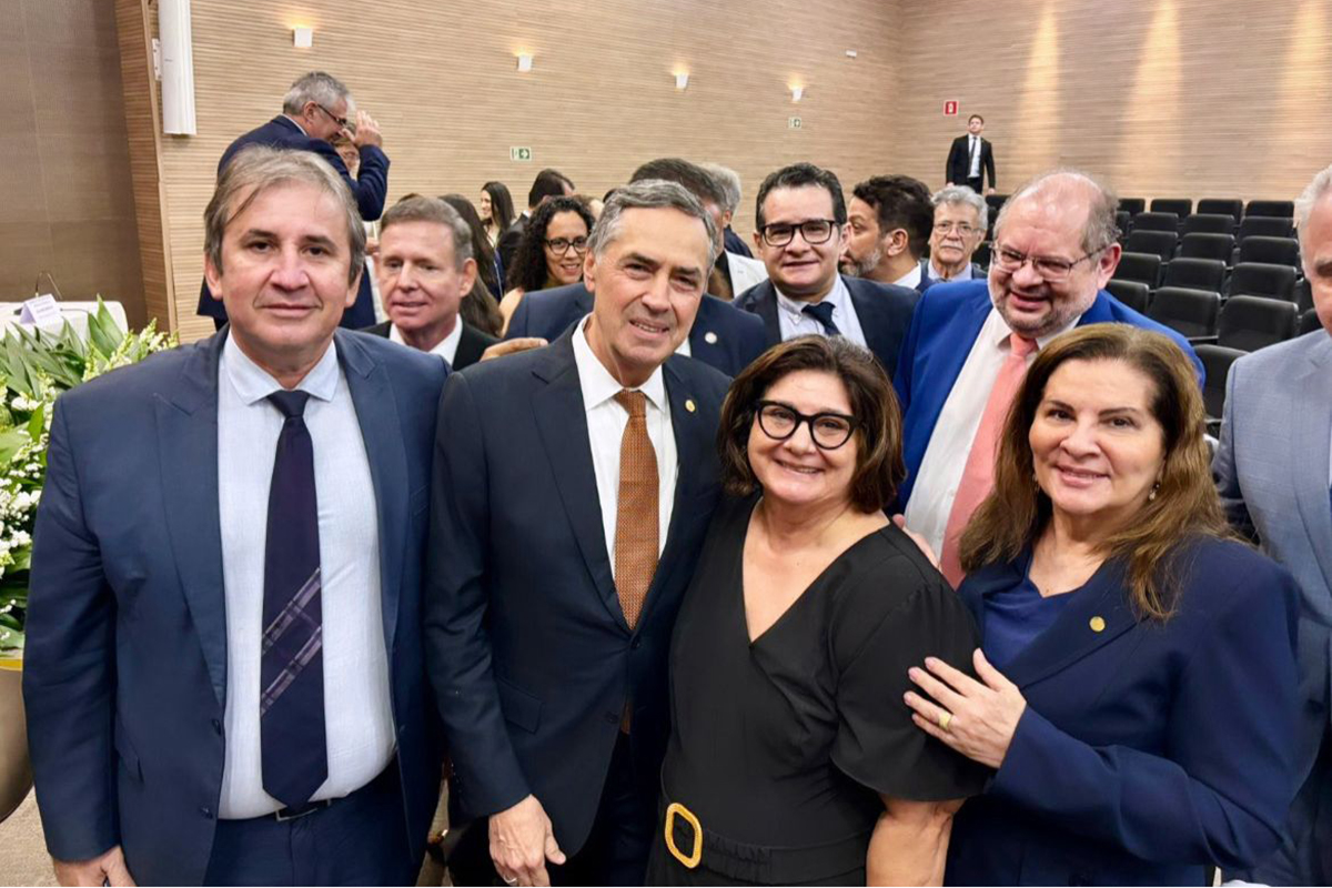 Em um evento, um grupo de sete pessoas, incluindo quatro homens de terno e três mulheres, sorri para a câmera. Ao fundo, um auditório com mais pessoas sugere uma ocasião formal. Todos parecem felizes e engajados.