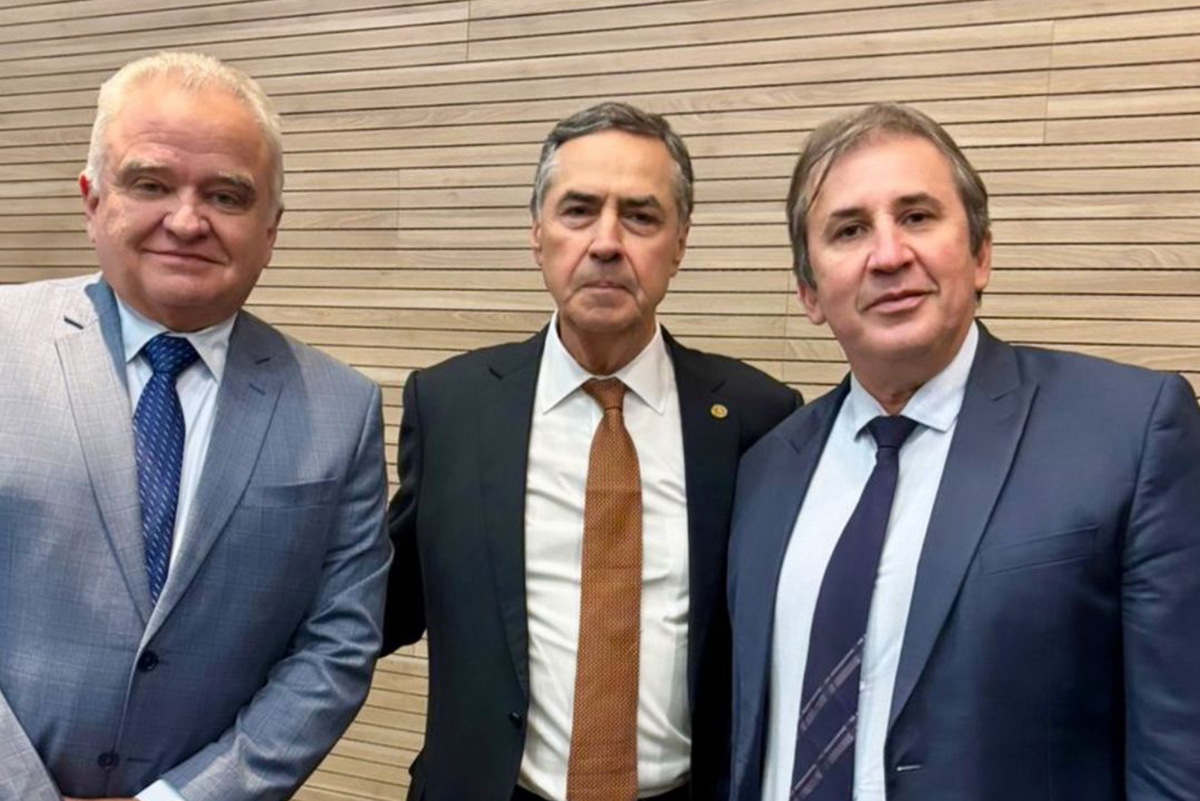 Três homens, todos de terno, posam lado a lado contra uma parede de madeira clara. O homem do centro veste um terno escuro com gravata marrom. Os outros dois vestem ternos azuis. Todos olham diretamente para a câmera.