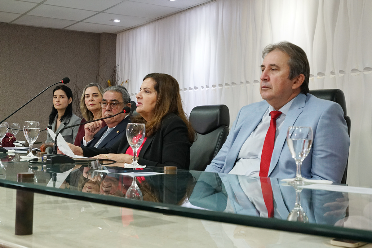  Cinco pessoas estão sentadas a uma longa mesa de vidro, participando de uma reunião. À direita, um homem de terno azul claro e gravata vermelha olha para a frente. As outras pessoas, incluindo três mulheres e um homem, estão atentas, algumas com papéis e copos na mesa.