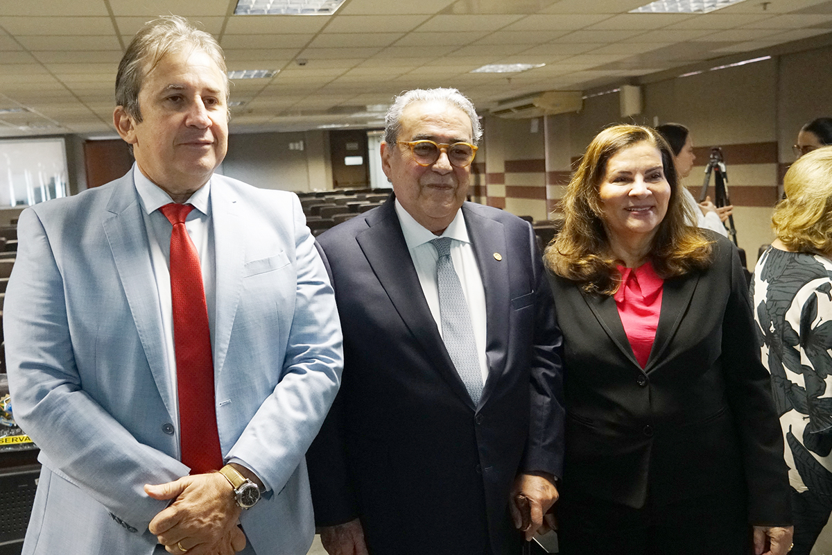 Três pessoas posam para uma foto: um homem de terno azul claro e gravata vermelha à esquerda, um homem mais velho de terno escuro no centro, e uma mulher de blazer preto e blusa rosa à direita. Eles sorriem para a câmera em um ambiente interno.