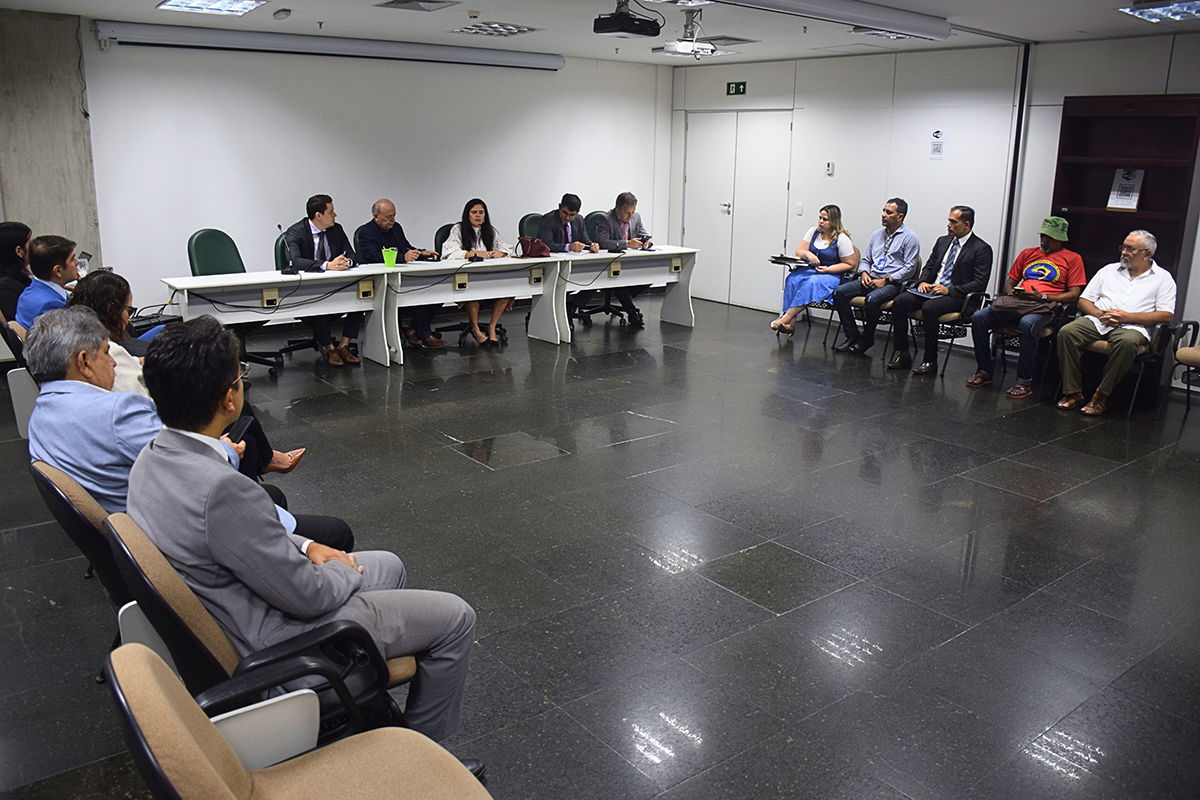 Uma visão mais ampla do mesmo ambiente revela o arranjo completo da sala. As cinco pessoas da imagem anterior estão sentadas à mesa principal, enquanto um grupo maior de participantes senta-se em cadeiras voltadas para a mesa, observando os palestrantes.