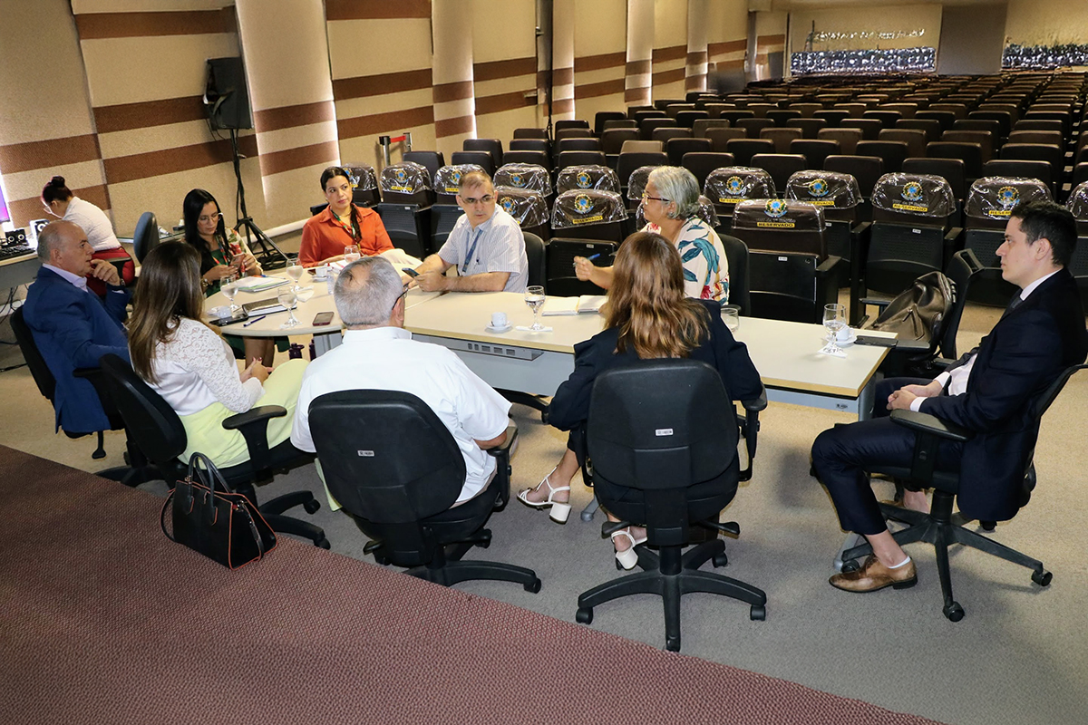 Um grupo de oito pessoas sentadas ao redor de uma mesa curva em uma sala grande com cadeiras vazias ao fundo. Quatro pessoas estão de costas para a câmera, outras quatro de frente, conversando animadamente. Copos e notebooks estão sobre a mesa.