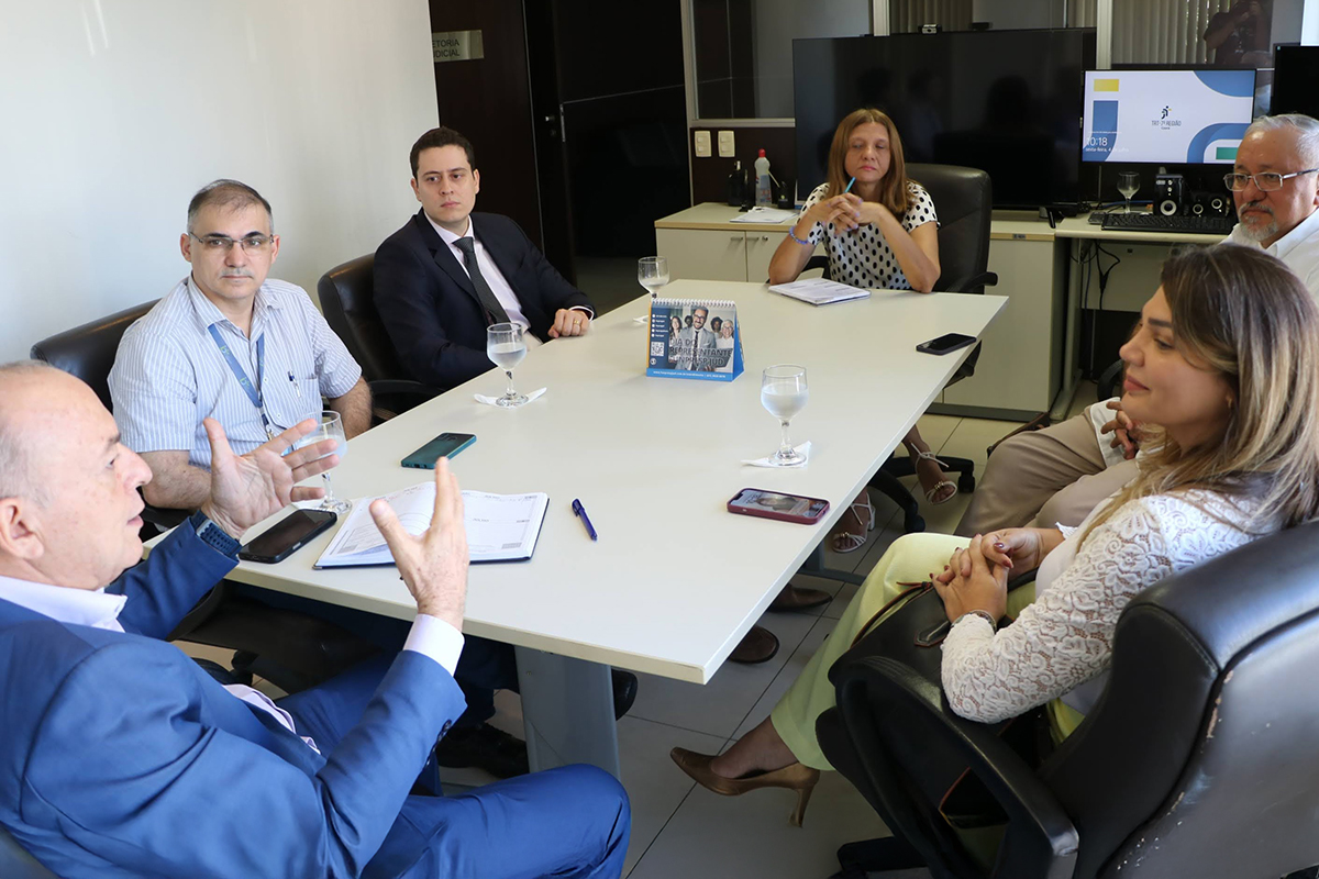 Uma reunião acontece em uma sala clara. Seis pessoas estão sentadas ao redor de uma mesa retangular branca. Duas pessoas à esquerda, uma à direita, e uma ao fundo. Um homem de terno azul gesticula enquanto fala, o foco em suas mãos. Copos e celulares estão sobre a mesa.