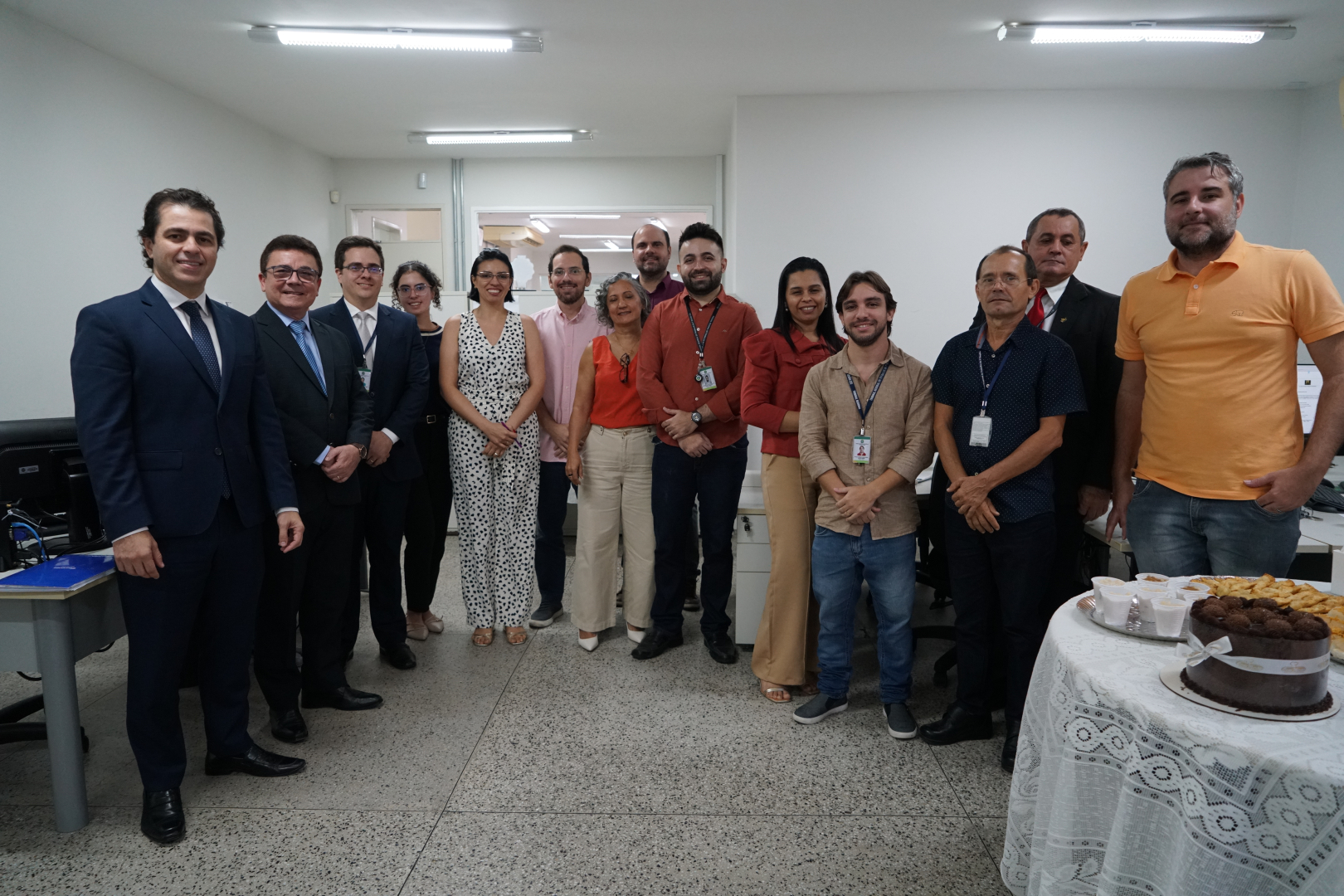 Fotografia colorida de um grupo de aproximadamente quinze pessoas em um ambiente de escritório. Homens e mulheres vestidos com roupas de trabalho posam juntos, de pé. No canto direito, em primeiro plano, há uma mesa redonda com uma toalha de renda branca, sobre a qual estão um bolo de chocolate e salgadinhos. Ao fundo, veem-se mesas e equipamentos de escritório, indicando uma celebração no local de trabalho.