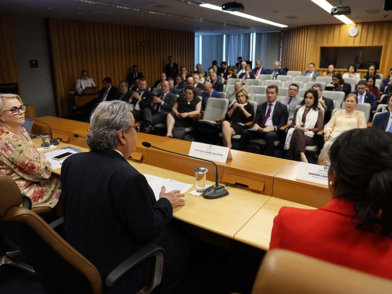 Pessoas estão sentadas em auditório em um evento, com a maioria olhando para um ponto central. O ministro Aloysio Corrêa de costas para a câmera, discursa com papéis nas mãos. Ao seu lado, à esquerda, uma mulher de cabelos loiros e óculos olha para ele.