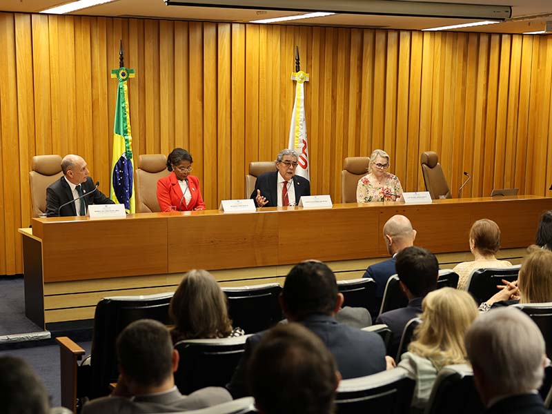 Quatro pessoas estão sentadas em uma mesa de madeira em um auditório, com duas bandeiras verticais atrás delas. O ministro Aloysio Corrêa está no centro, de terno e gravata vermelha, com os braços abertos gesticulando. Uma plateia parcialmente visível assiste ao evento, enquanto ele profere seu discurso.