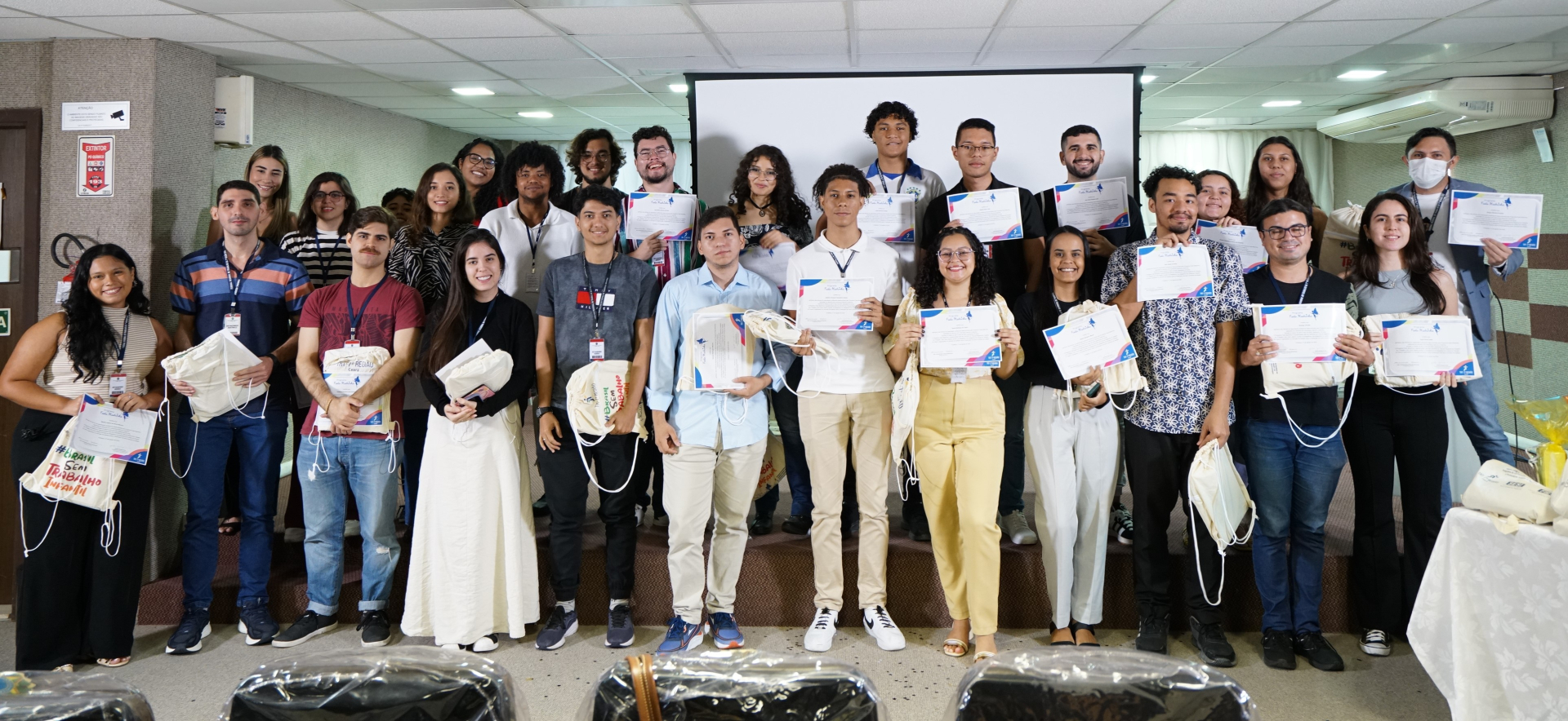 Foto de dezenas de estagiários exibindo certificados de conclusão das oficinas do Programa Fada Madrinha. Eles estão em pé em cima do palco do auditório da Escola Judicial. Estão sorridentes e posando para a foto
