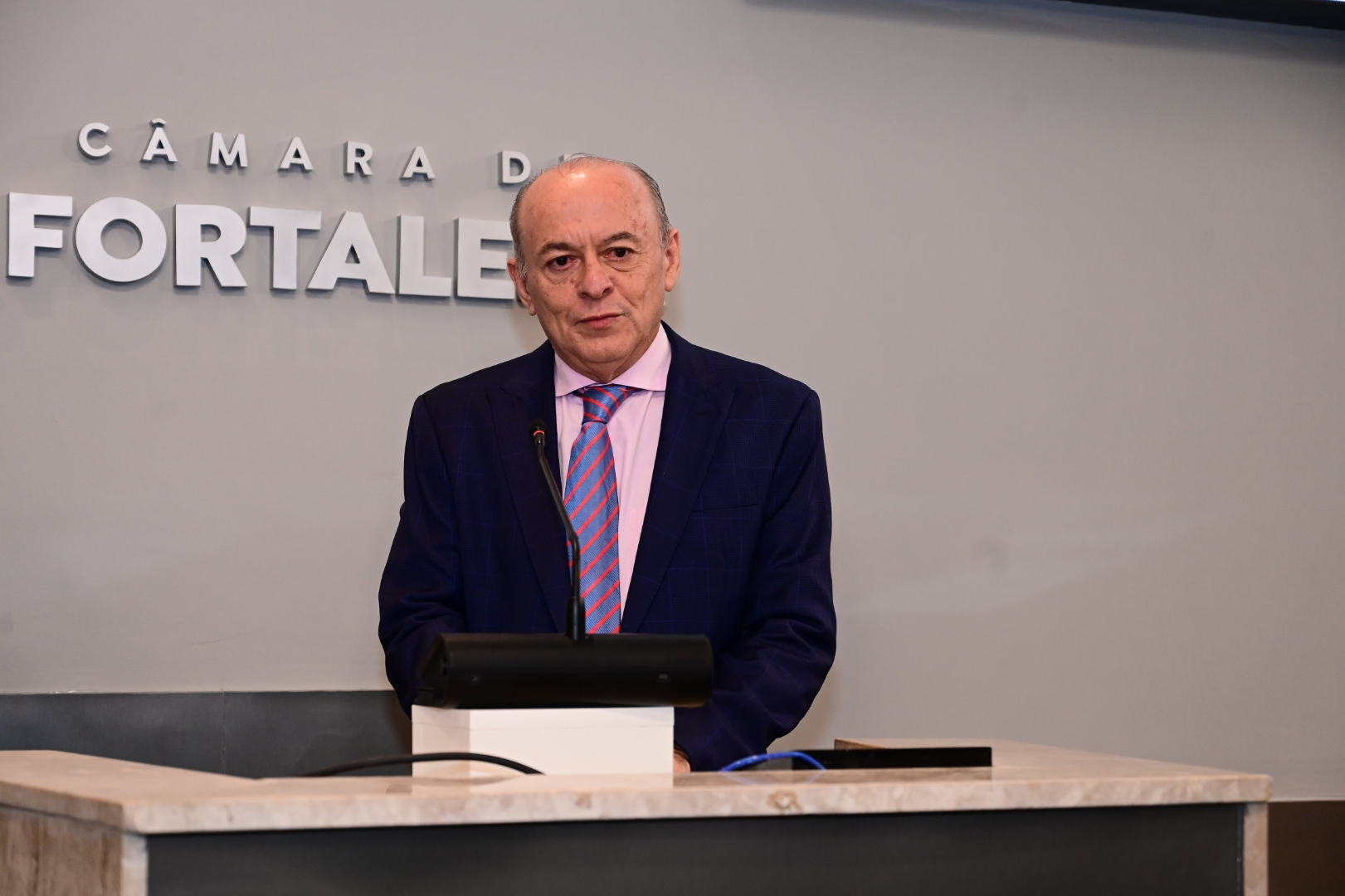 Um homem de meia-idade (des. Parente), calvo e com uma expressão séria, está em pé atrás de um púlpito com tampo de mármore. Ele veste um terno azul-marinho com um colete azul, camisa branca e uma gravata rosa listrada. No fundo, uma parede cinza exibe em relevo as palavras "CÂMARA DE FORTALEZA".