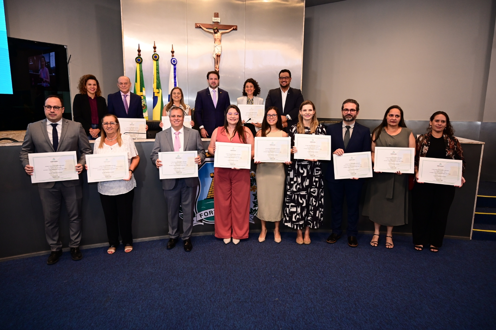Um grupo de 15 pessoas, composto por homens e mulheres, está em pé em duas filas em um auditório. Todos seguram certificados emoldurados, exibindo-os para a câmera. Ao fundo, uma parede cinza tem um crucifixo central e bandeiras do Brasil, Ceará e Fortaleza. Também há a imagem de um Cristo no fundo da parede.