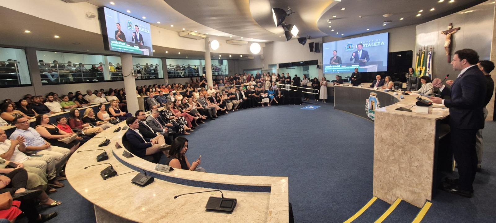  Vista panorâmica de um auditório lotado, com uma plateia sentada em assentos semicirculares azuis. No centro, uma mesa de honra elevada com autoridades, onde um homem de terno fala ao microfone. No fundo, há telões exibindo o logotipo da Câmara Municipal de Fortaleza.