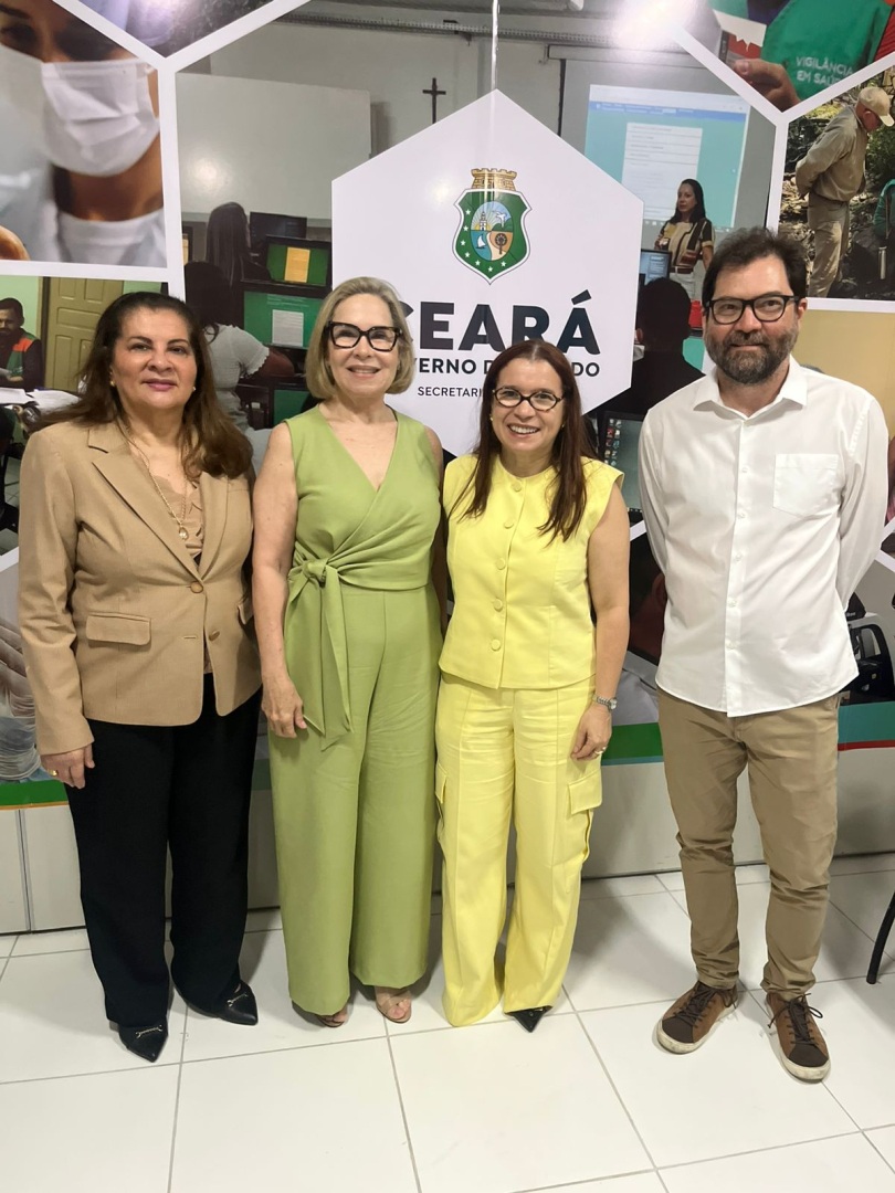 A imagem mostra quatro pessoas de pé, alinhadas em frente a uma parede de fundo com várias fotos e no centro o logotipo do Governo do Estado do Ceará. Da esquerda para a direita, uma mulher de blazer bege e calça preta, uma mulher com um macacão verde, uma mulher vestida de amarelo, e um homem de camisa branca e calça bege. Todos sorriem para a câmera.