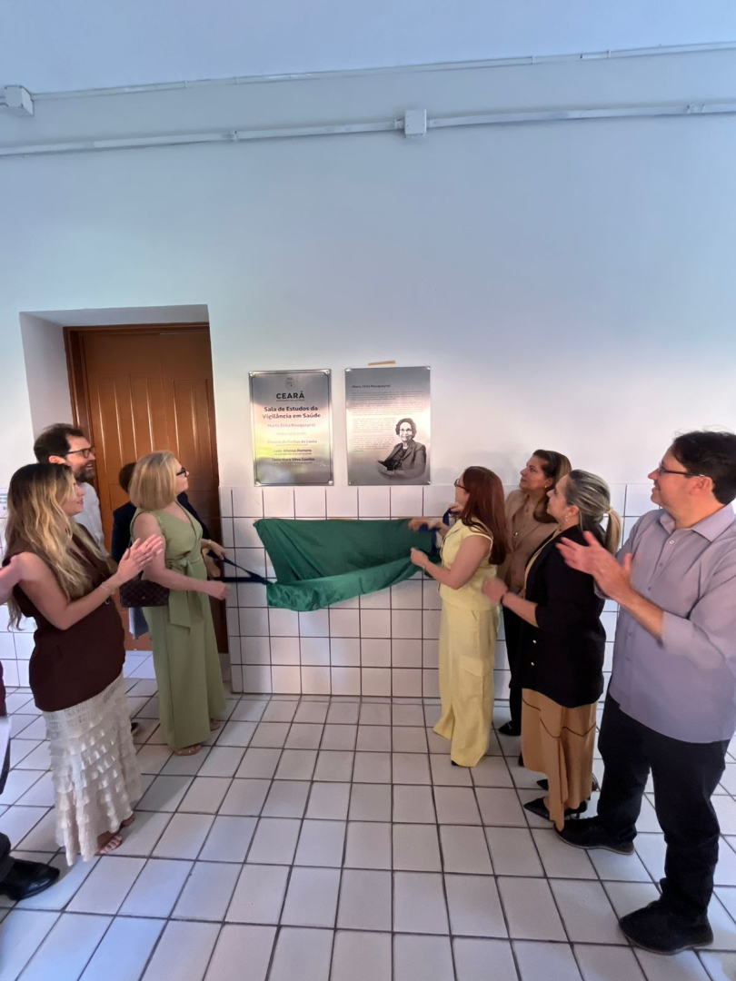 A imagem registra a inauguração de uma placa em uma parede branca. Várias pessoas, em trajes casuais, estão ao redor da placa, aplaudindo e sorrindo. Uma mulher de vestido amarelo e outra com roupa preta e calça bege estão puxando o pano verde que cobria a placa, revelando-a. As placas são de metal prateado e têm inscrições e a foto de uma pessoa.