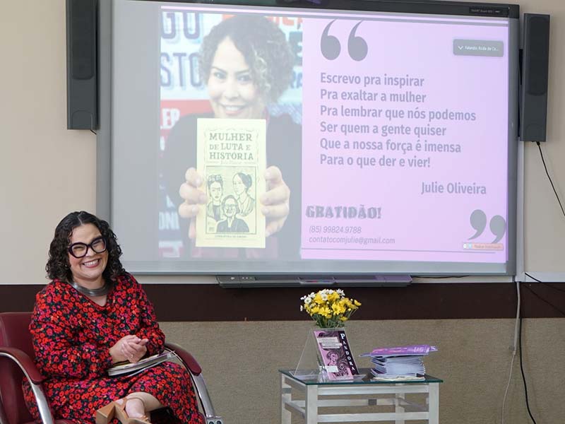 Uma mulher de cabelo escuro cacheado, óculos e vestido estampado vermelho senta em uma poltrona escura. Ela sorri enquanto segura um caderno fechado. Atrás dela, uma tela projeta uma imagem com sua foto e uma citação