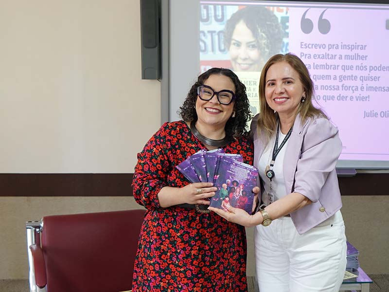 Duas mulheres sorriem para a câmera. A da esquerda, com cabelo cacheado e vestido florido vermelho, segura uma pilha de livros de capa roxa. A da direita, de blazer lilás e calça branca, aponta para os livros. Ao fundo, uma projeção mostra um rosto feminino e uma citação