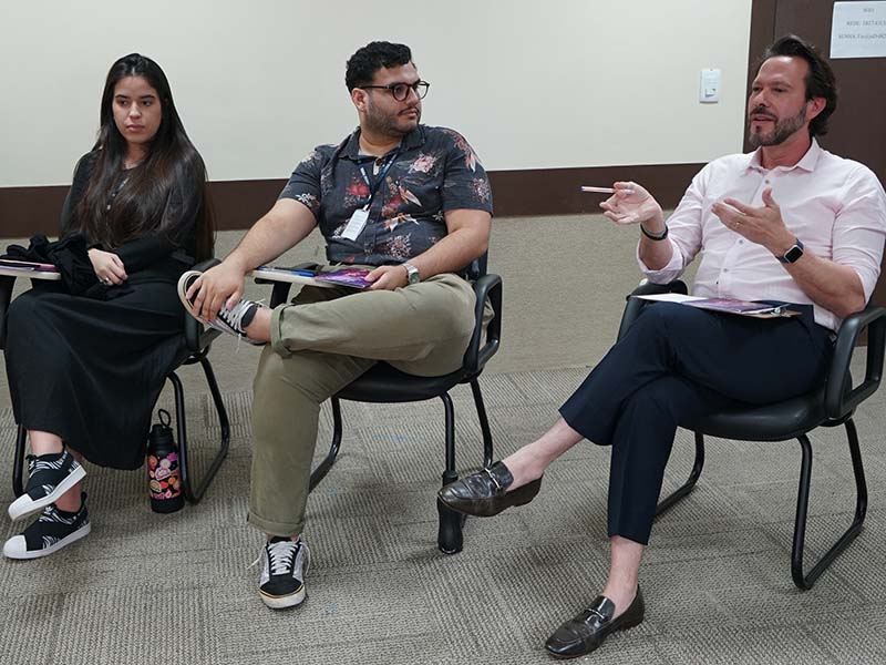 Três pessoas sentadas em cadeiras, da esquerda para a direita: uma mulher de cabelo escuro comprido e vestido preto, um homem barbudo de camisa florida e calça cáqui, e um homem de camisa social rosa claro e calça azul escura, gesticulando enquanto fala.